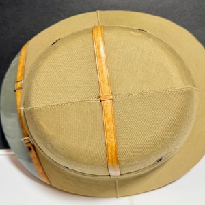 May include: A tan pith helmet featuring a rounded crown and a wide brim. The exterior is accented with a vertical tan leather strap running over the top and a horizontal leather band around the base, secured with a small silver-toned metal buckle.