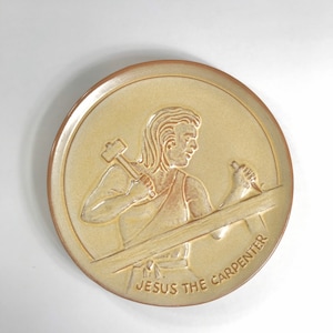 Puede incluir: Plato redondo de cerámica beige con un diseño en relieve. El diseño representa una figura con un martillo y madera, con el texto "JESUS THE CARPENTER". El plato tiene un borde marrón.