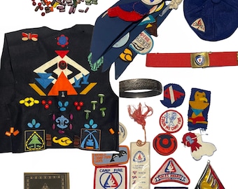 Op de afbeelding: Een verzameling vintage Girl Scout camp-patches en -badges. De patches zijn voornamelijk blauw, rood en geel met verschillende ontwerpen, waaronder een kampvuur, een zeilboot en een berg. Er zijn ook wat kralen en een boek met de titel "The Shulutamna of the Camp Fire Girls".