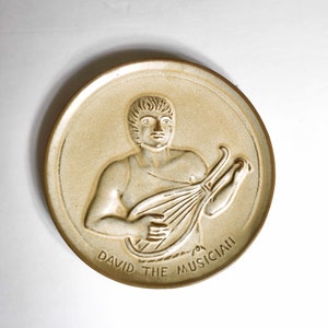 Puede incluir: Un plato de cerámica beige con una imagen en relieve de un hombre tocando una lira. El plato está inscrito con las palabras "David the Musician".