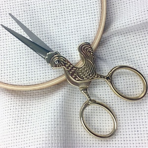 May include: Gold-toned embroidery scissors with a rooster design. The scissors are lying on a white Aida cloth with a wooden embroidery hoop.
