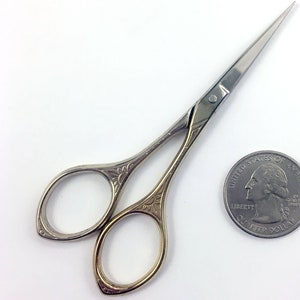 May include: A pair of small, silver-toned scissors with ornate floral designs on the handles. The scissors are about the size of a UK 20 pence coin.