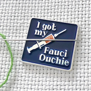 Op de afbeelding: Een blauwe en zilveren emaille pin met een spuit en de tekst "I got my Fauci Ouchie".