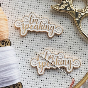 May include: Two gold-colored enamel pins with white backgrounds. The pins say "I'm speaking" in a cursive font. The pins are on a white fabric background with a wooden embroidery hoop and a pair of gold scissors.