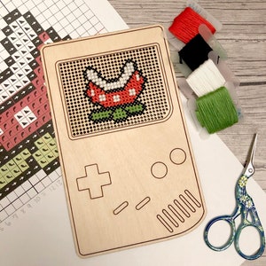 May include: A wooden craft kit featuring a pixelated image of a Piranha Plant from the Super Mario Bros. video game. The kit includes embroidery floss in red, black, white, and green, a pair of scissors, and a wooden frame.