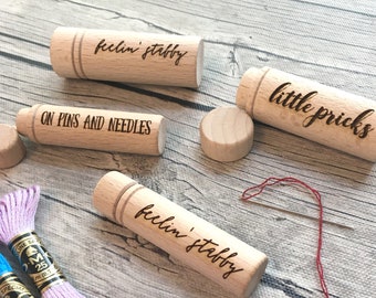 May include: Three wooden needle cases with the text "feelin' stabby" and "little pricks" on them. One case has the text "ON PINS AND NEEDLES". The cases are on a wooden surface with spools of embroidery floss.