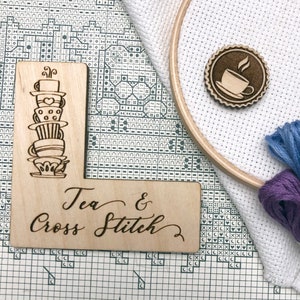 May include: Wooden sign with the text "Tea & Cross Stitch" and a drawing of a stack of teacups. The sign is on a white Aida cloth with a wooden embroidery hoop and purple and blue embroidery floss.