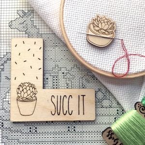 K&ouml;nnte beinhalten: Ein Holz-Bastelset mit zwei Sukkulenten-Designs. Das Set enth&auml;lt ein St&uuml;ck Holz mit dem Text "SUCC IT" und eine kleine Holz-Sukkulentenpflanze. Das Set befindet sich auf einem wei&szlig;en und schwarzen Gitterhintergrund.