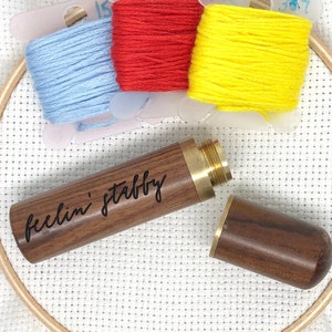 Pode incluir: Um porta-agulhas de madeira com o texto "feelin' stabby" gravado nele. O porta-agulhas está aberto e a metade superior está separada da metade inferior. O porta-agulhas está sobre um tecido Aida branco com três novelos de fio de bordado no fundo. O fio é azul, vermelho e amarelo.