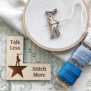 May include: Two wooden signs with the text "Talk Less Stitch More" and a silhouette of a person holding a needle. The signs are on a white background with a cross-stitch pattern.