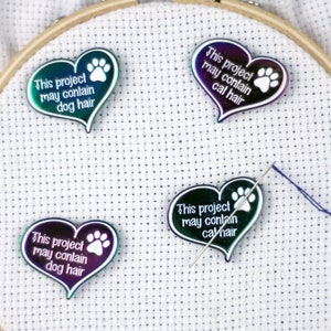 May include: Four iridescent heart-shaped pins with paw prints and the text "This project may contain dog hair" or "This project may contain cat hair". The pins are on a white fabric background.
