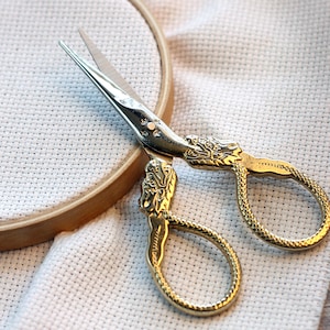 May include: Gold embroidery scissors with ornate handles on a white Aida cloth with a wooden embroidery hoop.