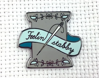 May include: A grey and blue enamel pin featuring a spool of thread with a needle and the text "Feelin' stabby".