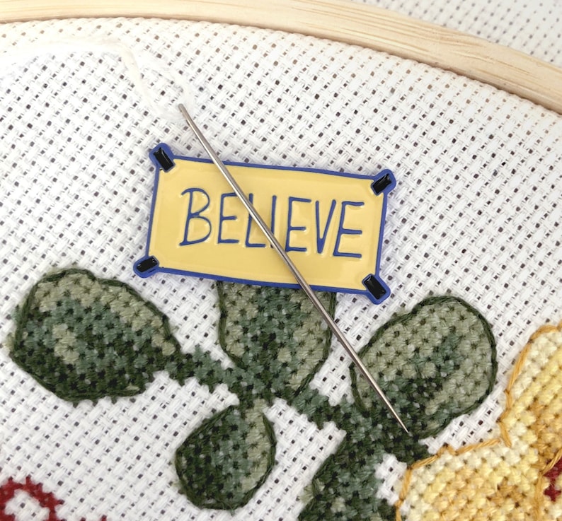 K&ouml;nnte beinhalten: Nahaufnahme eines Kreuzstichprojekts mit einem gelben Banner, auf dem "BELIEVE" steht, und einer Nadel und einem Faden, die durch den Stoff gehen.
