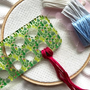May include: A cross-stitch project in progress, featuring a wooden embroidery hoop with white fabric. A clear plastic template with a cactus pattern is positioned near a knot of red thread. Spools of white, gray, and blue embroidery floss are also visible.