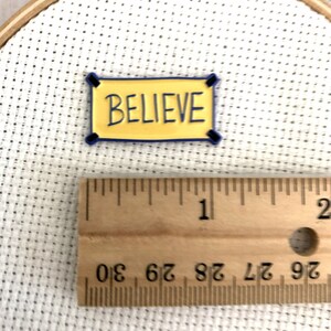 K&ouml;nnte beinhalten: Ein gelbes Banner mit blauem Besatz und dem Wort "BELIEVE" in blauen Buchstaben. Das Banner ist etwa 2,5 cm breit und liegt auf einer wei&szlig;en Oberfl&auml;che.