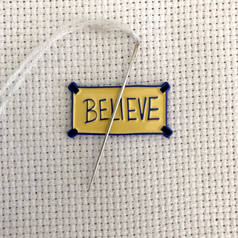 K&ouml;nnte beinhalten: Ein wei&szlig;er Faden ist in eine Nadel eingef&auml;delt, die &uuml;ber einem gelben Banner mit dem Wort "BELIEVE" in blauer Schrift platziert ist. Das Banner hat einen blauen Rand und befindet sich auf einem wei&szlig;en Aida-Stoff.