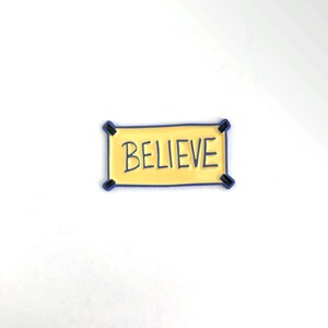 K&ouml;nnte beinhalten: Eine gelbe Emaille-Anstecknadel mit blauem Rand, die das Wort "BELIEVE" in schwarzen Buchstaben zeigt. Die Anstecknadel hat die Form eines Banners mit zwei schwarzen Laschen oben.