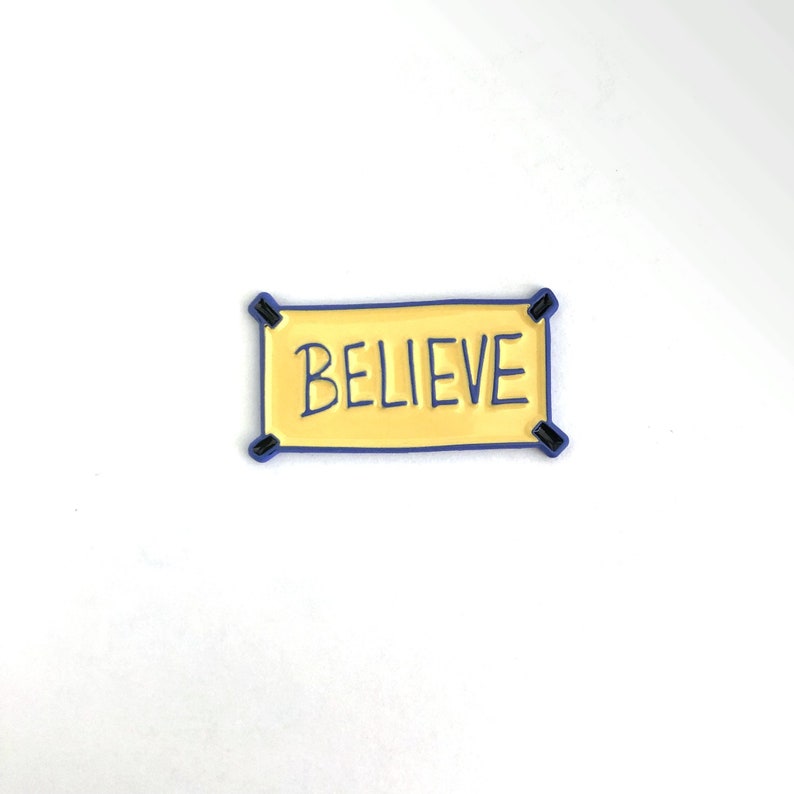 K&ouml;nnte beinhalten: Eine gelbe Emaille-Anstecknadel mit blauem Rand, die das Wort "BELIEVE" in schwarzen Buchstaben zeigt. Die Anstecknadel hat die Form eines Banners mit zwei schwarzen Laschen oben.