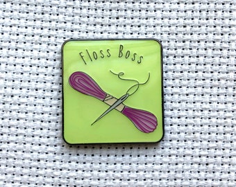 Può includere: Uno spillo in smalto verde con contorno nero. Lo spillo presenta un ago e filo con un rocchetto di filo viola. Il testo "Floss Boss" è scritto in nero sullo spillo.
