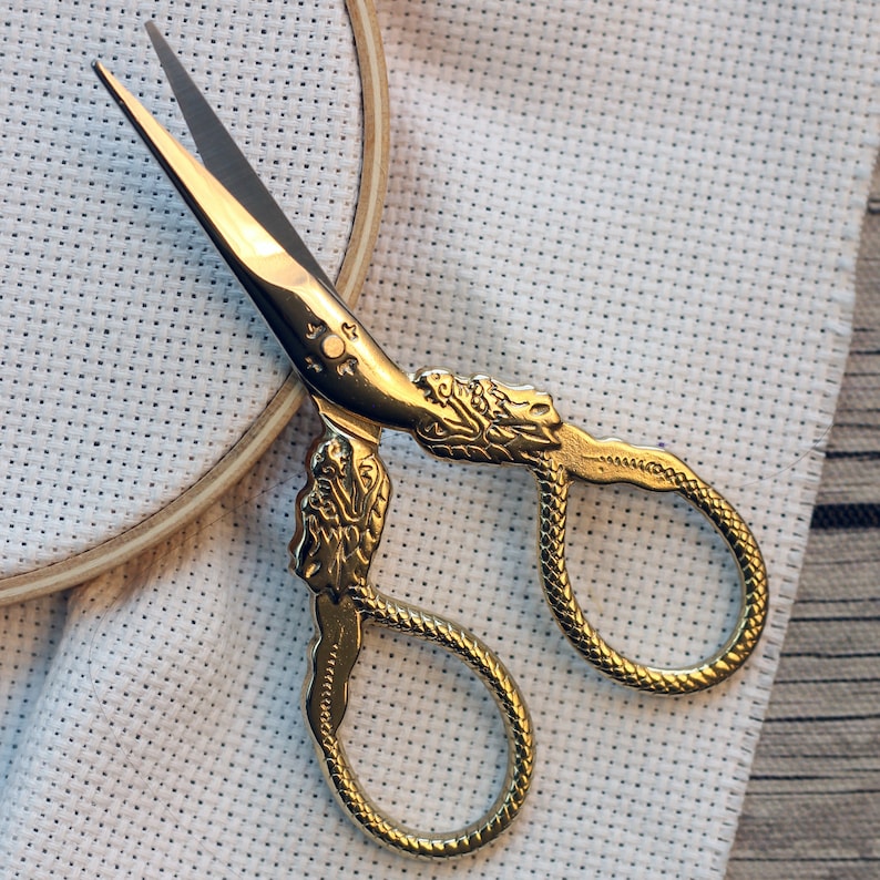 May include: Gold-toned embroidery scissors with ornate dragon-like handles. The scissors are lying on a white Aida cloth.