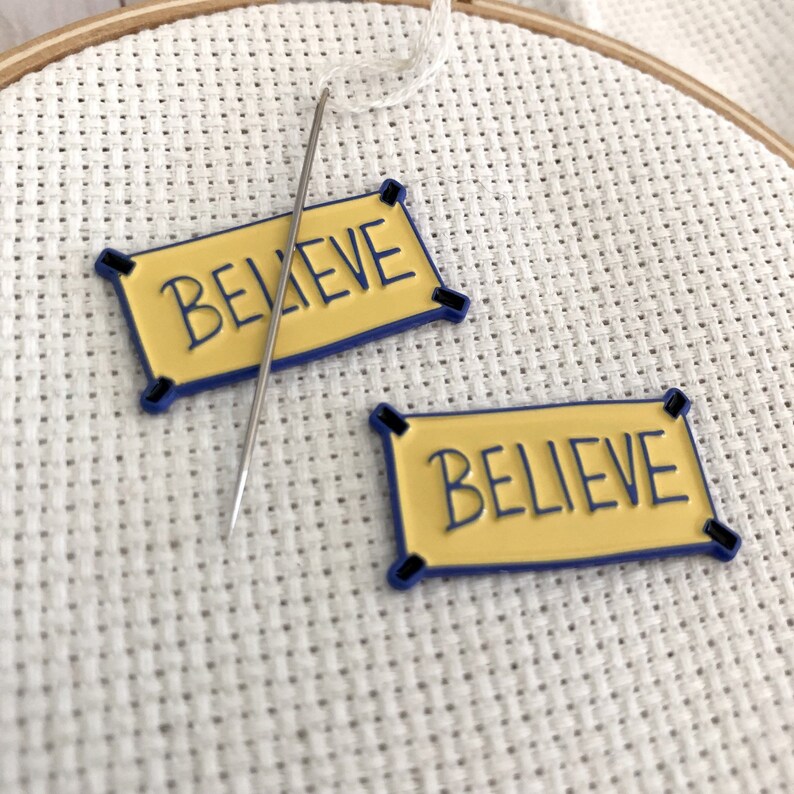 K&ouml;nnte beinhalten: Zwei gelbe Filzbanner mit blauem Besatz und schwarzen N&auml;hten, jeweils mit dem Wort "BELIEVE" in Schwarz geschrieben. Eine Nadel und wei&szlig;er Faden sind im Hintergrund sichtbar.