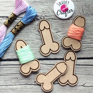 Peut inclure: Quatre porte-fils en bois en forme d'os avec du fil à broder de différentes couleurs enroulé autour. Les supports sont sur un fond en bois clair. Le texte "SNARKY CRAFTER" est sur un bouton rond.