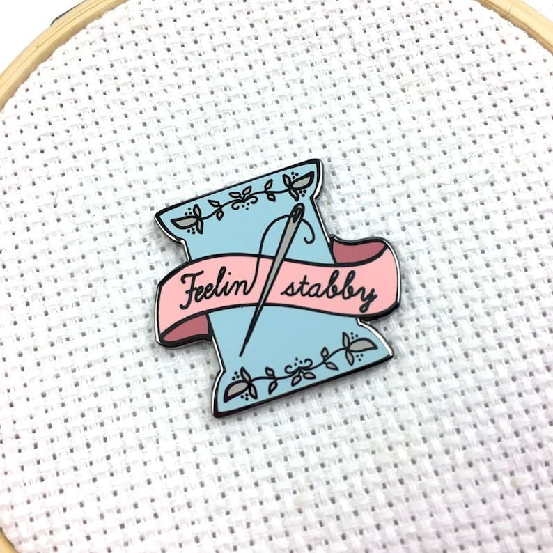 May include: A blue and pink enamel pin with a needle and thread in a spool and a banner that reads "Feelin' stabby".