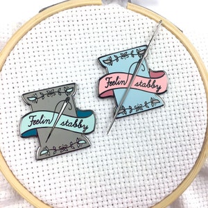 May include: Two enamel pins shaped like spools of thread with needles sticking out. The spools are in shades of blue and grey. The text on the banners reads "Feelin' stabby".