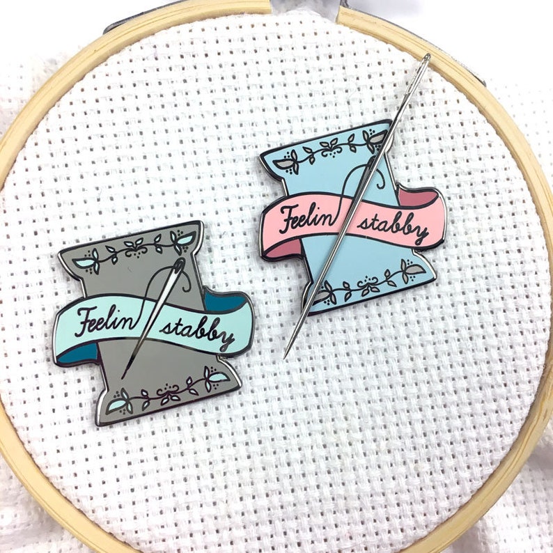May include: Two enamel pins shaped like spools of thread with needles sticking out. The spools are in shades of blue and grey. The text on the banners reads "Feelin' stabby".