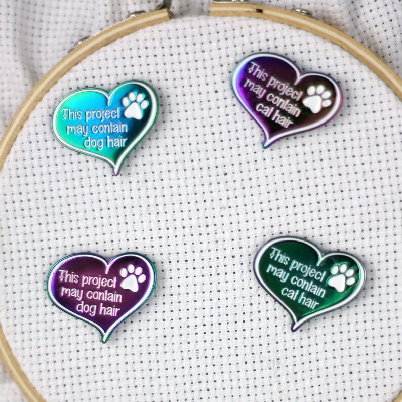 May include: Four iridescent heart-shaped pins with white paw prints and text that reads "This project may contain dog hair" and "This project may contain cat hair".