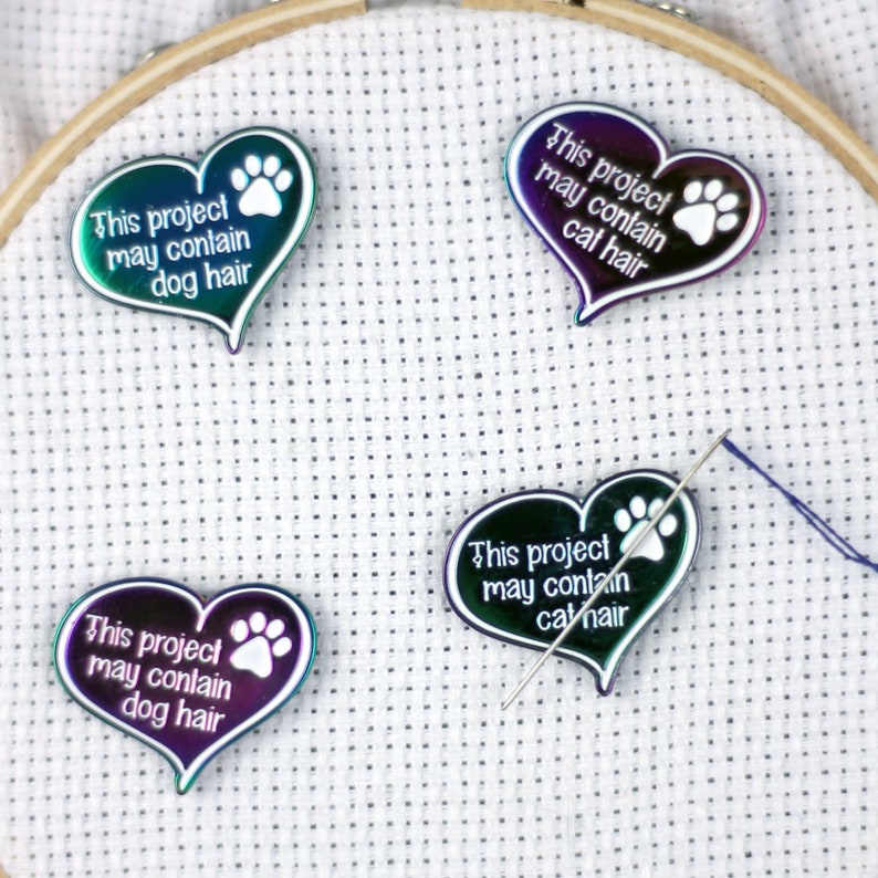 May include: Four iridescent heart-shaped pins with paw prints and the text "This project may contain dog hair" or "This project may contain cat hair". The pins are on a white fabric background.
