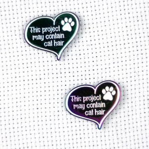 May include: Two iridescent heart-shaped metal pins with a white paw print and the text "This project may contain cat hair".