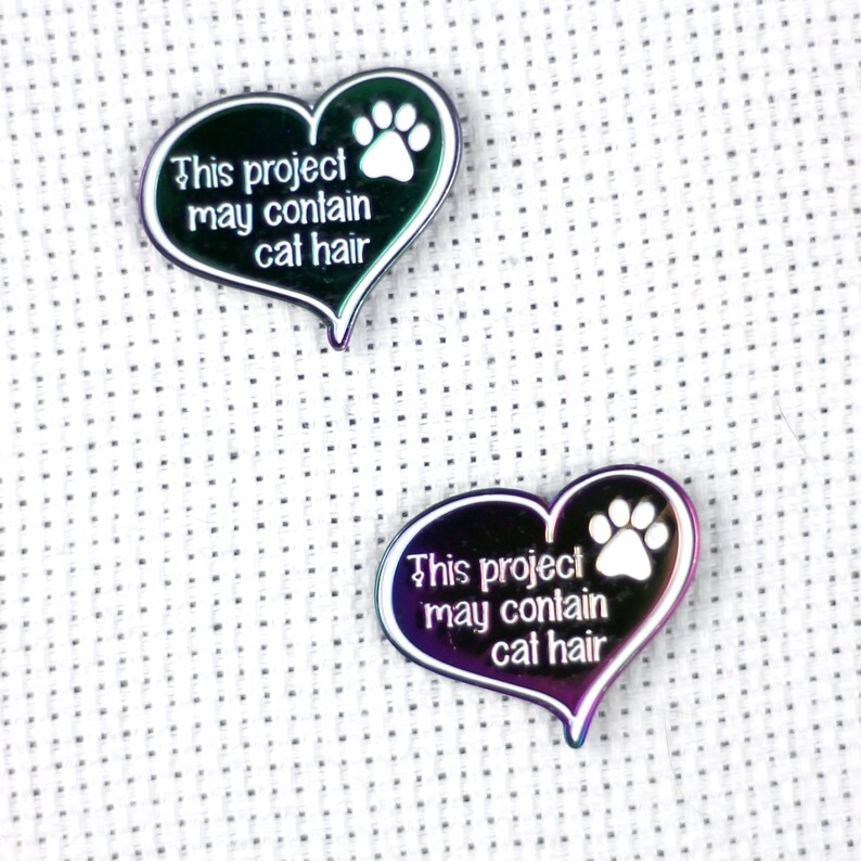 May include: Two iridescent heart-shaped metal pins with a white paw print and the text "This project may contain cat hair".