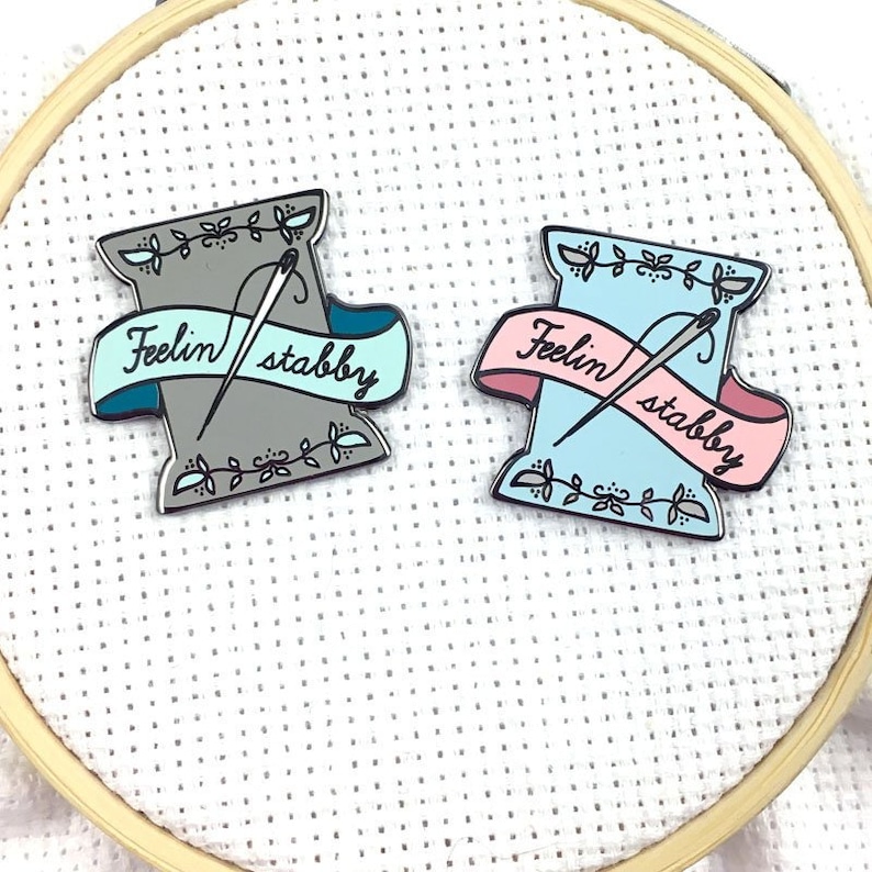May include: Two enamel pins shaped like a spool of thread with a needle and the text "Feelin' stabby". One pin is grey with a teal banner and the other is blue with a pink banner.