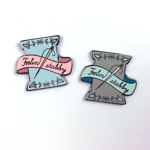 May include: Two enamel pins shaped like spools of thread with needles and the text "Feelin' stabby" on a banner. One pin is light blue and the other is grey.