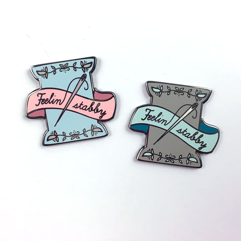 May include: Two enamel pins shaped like spools of thread with needles and the text "Feelin' stabby" on a banner. One pin is light blue and the other is grey.