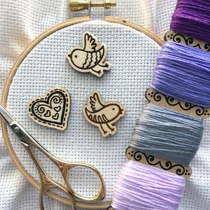 Könnte beinhalten: Ein weißer Stickrahmen mit einem Herz und zwei Vögeln, alles aus Holz. Der Rahmen ist von Spulen mit violettem, lavendelfarbenem, grauem und hellviolettem Stickgarn umgeben.