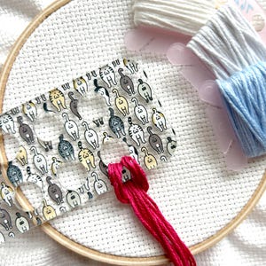 May include: A cross-stitch project in progress, featuring a wooden embroidery hoop and white fabric. A clear plastic floss organizer with a cat butt pattern is visible. Pink, white, light blue, and gray embroidery floss are also present.