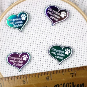 May include: Four iridescent heart-shaped pins with paw prints and the text "This project may contain [animal] hair" in white. The pins are arranged on a white fabric background.
