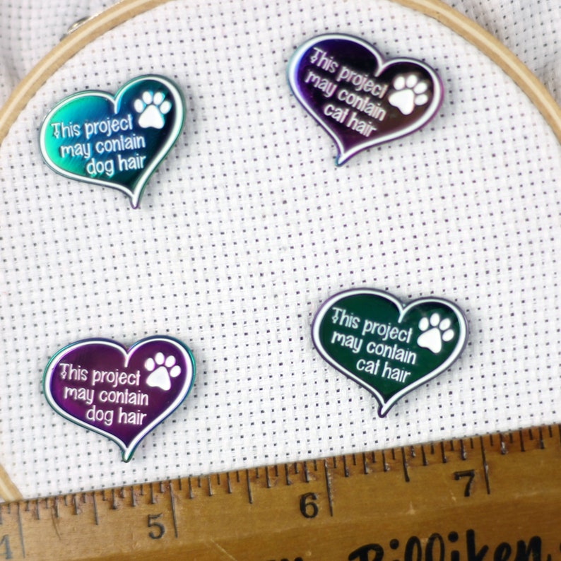 May include: Four iridescent heart-shaped pins with paw prints and the text "This project may contain [animal] hair" in white. The pins are arranged on a white fabric background.