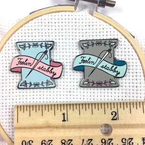 May include: Two enamel pins shaped like spools of thread with needles and a banner that reads "Feelin' stabby". One pin is blue and pink, the other is grey and teal. The pins are displayed on a white fabric background with a wooden ruler.