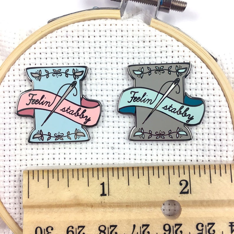 May include: Two enamel pins shaped like spools of thread with needles and a banner that reads "Feelin' stabby". One pin is blue and pink, the other is grey and teal. The pins are displayed on a white fabric background with a wooden ruler.