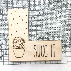 K&ouml;nnte beinhalten: Ein Holzschild mit einer Illustration einer Sukkulente und dem Text "SUCC IT" in handschriftlicher Schrift.
