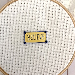 K&ouml;nnte beinhalten: Ein gelbes Banner mit blauem Rand und dem Wort "BELIEVE" in Schwarz.