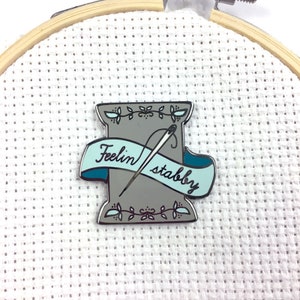 May include: A grey and blue enamel pin featuring a spool of thread with a needle and the text "Feelin' stabby".