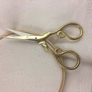 May include: Gold-toned embroidery scissors with a snake-like design on the handles. The scissors are lying on a white fabric surface.