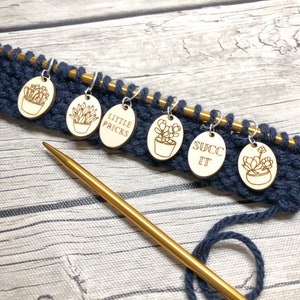 May include: Six wooden oval-shaped stitch markers with a succulent design and funny sayings. The markers are on a blue knitting project with a gold knitting needle.
