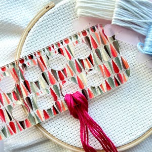 May include: A cross-stitch project in progress, featuring a white fabric stretched within a wooden embroidery hoop. A clear plastic tool with a colorful teardrop pattern is positioned over the fabric. Bright pink embroidery floss is threaded through the fabric, with spools of white and blue floss nearby.