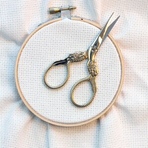 May include: A gold-colored embroidery scissors with ornate handles sits inside a wooden embroidery hoop on a white fabric background.